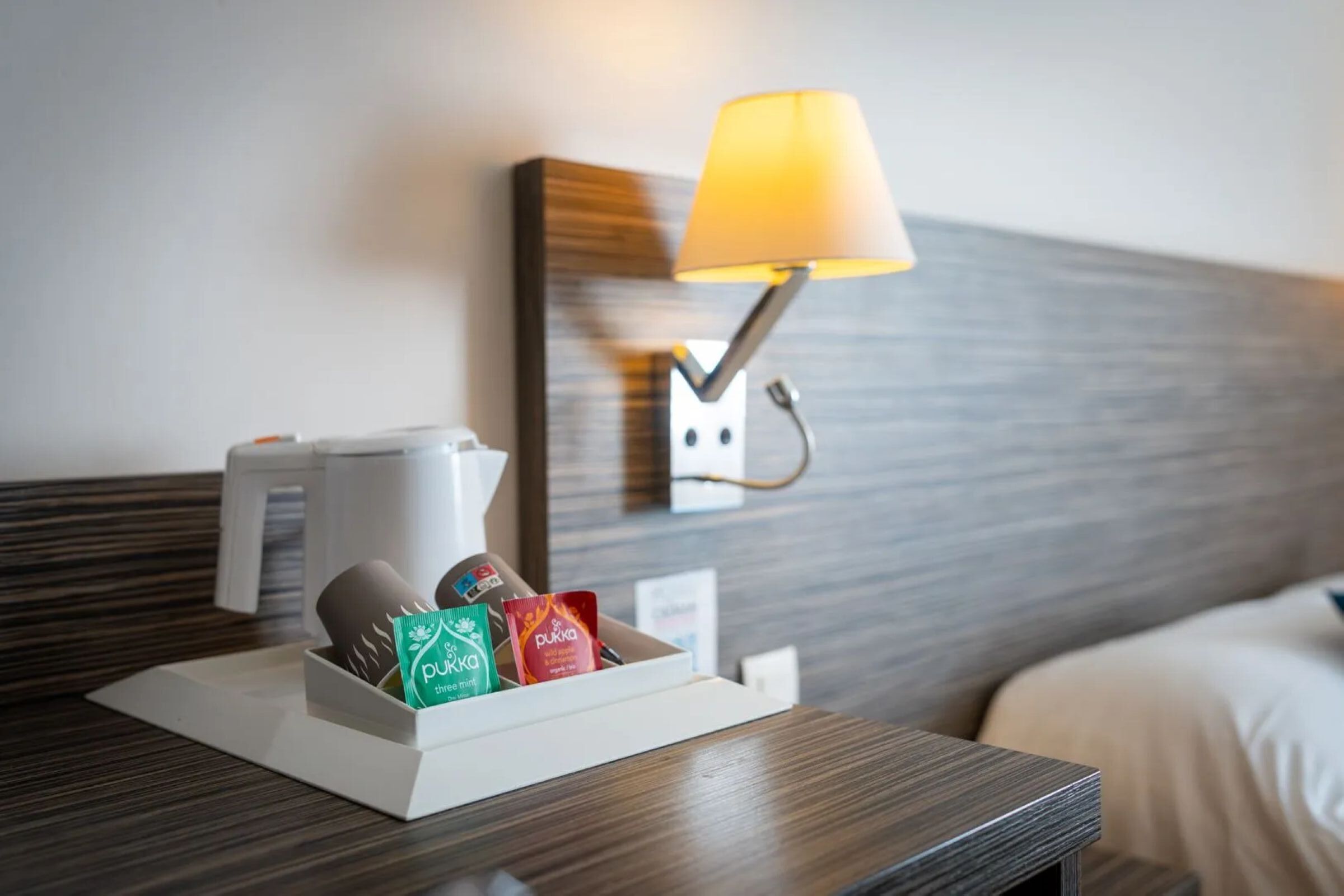 Una habitación de hotel con una mesita de noche que contiene una cafetera, una lámpara y una caja de bolsitas de té.