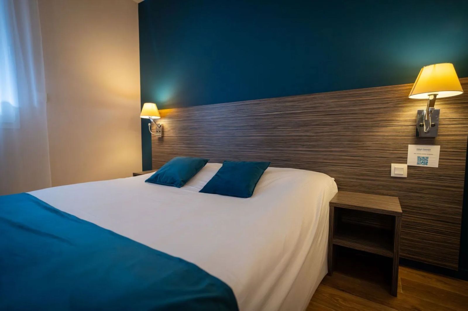 A hotel room with a large bed, two pillows, a nightstand, and wall-mounted lamps.