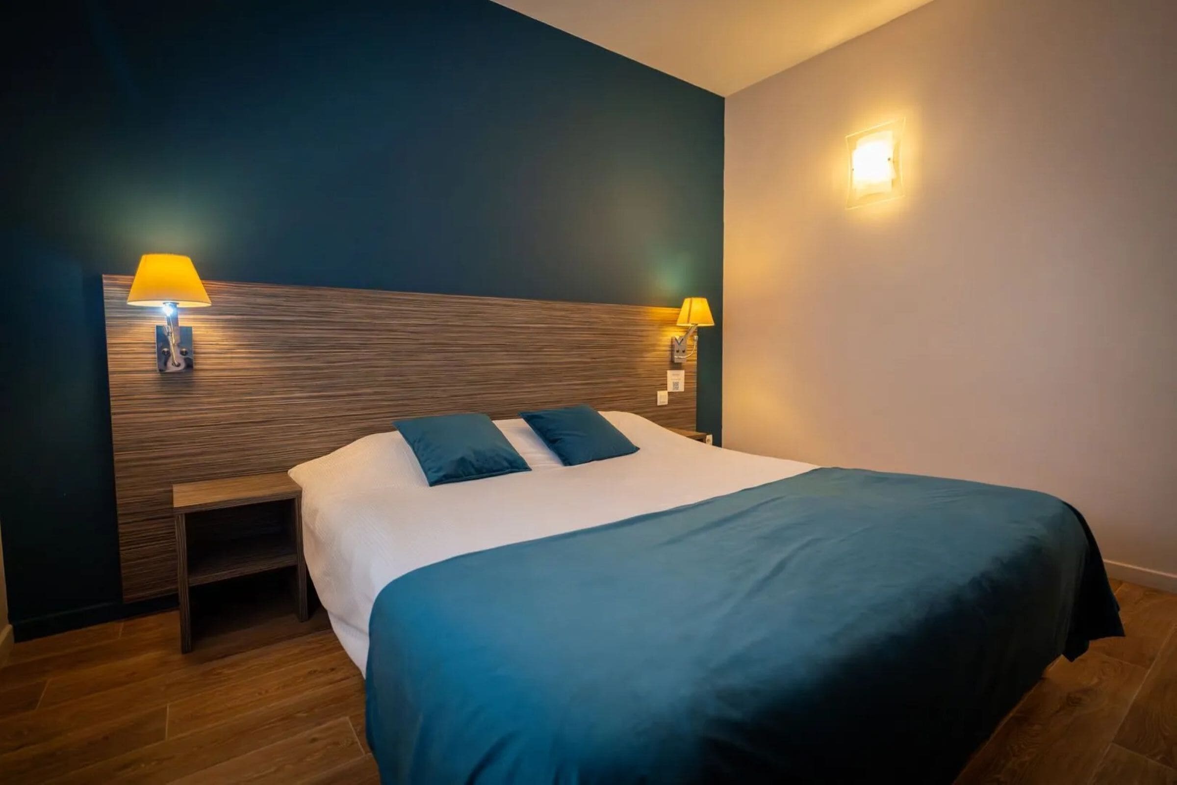 Una cama bien hecha con una colcha azul oscuro y dos almohadas, flanqueada por dos mesitas de noche con lámparas.