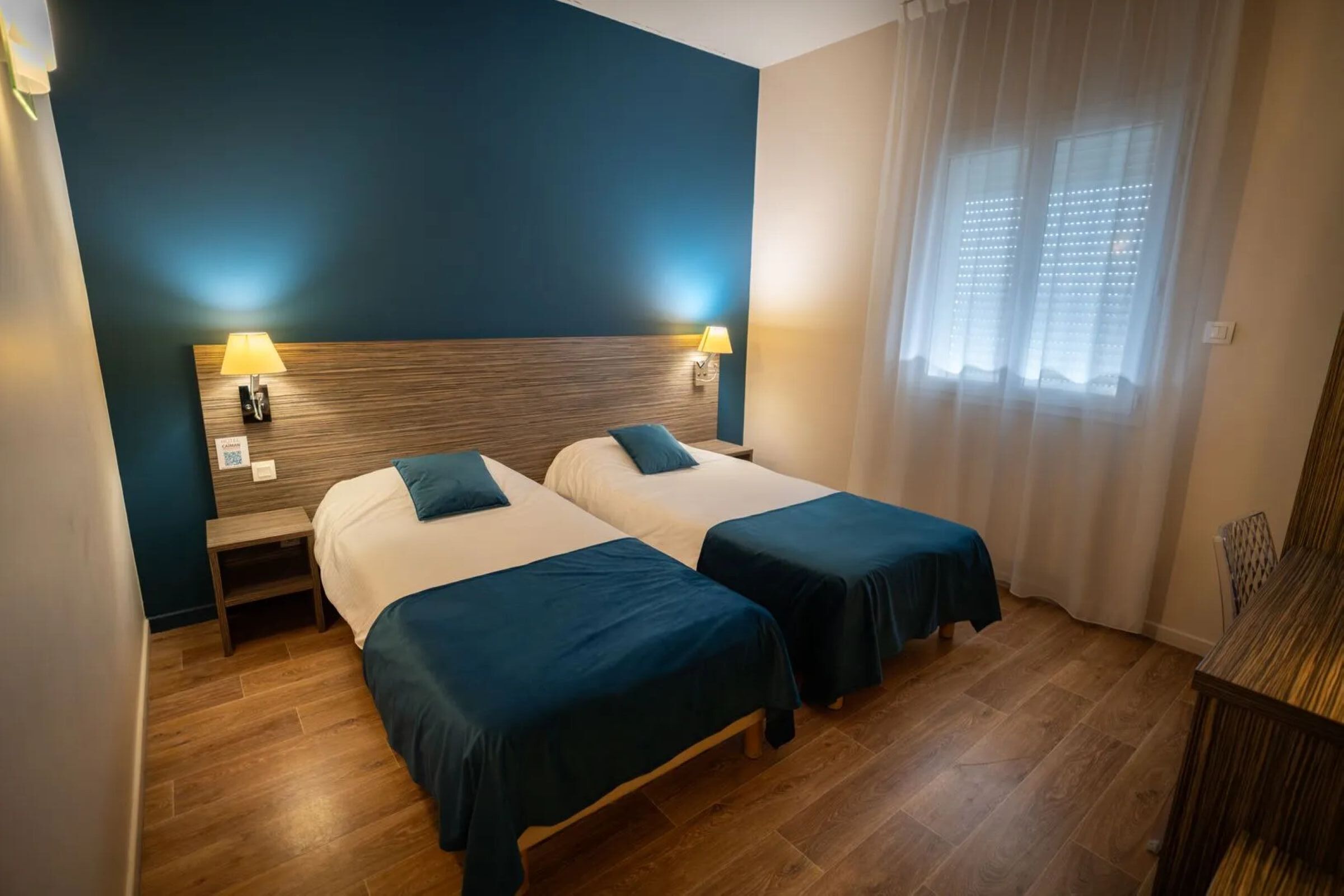 Habitación de hotel con dos camas, cabecero de madera, lámparas de noche y una ventana con cortinas translúcidas.