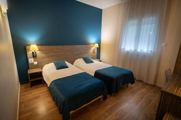 A hotel room with two beds, wooden headboard, bedside lamps, and a window with sheer curtains.