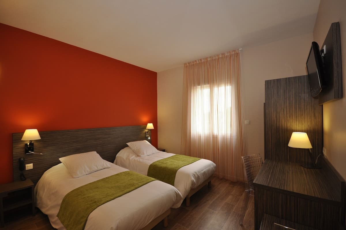 A hotel room with two beds, a desk, a TV, and a window with curtains.