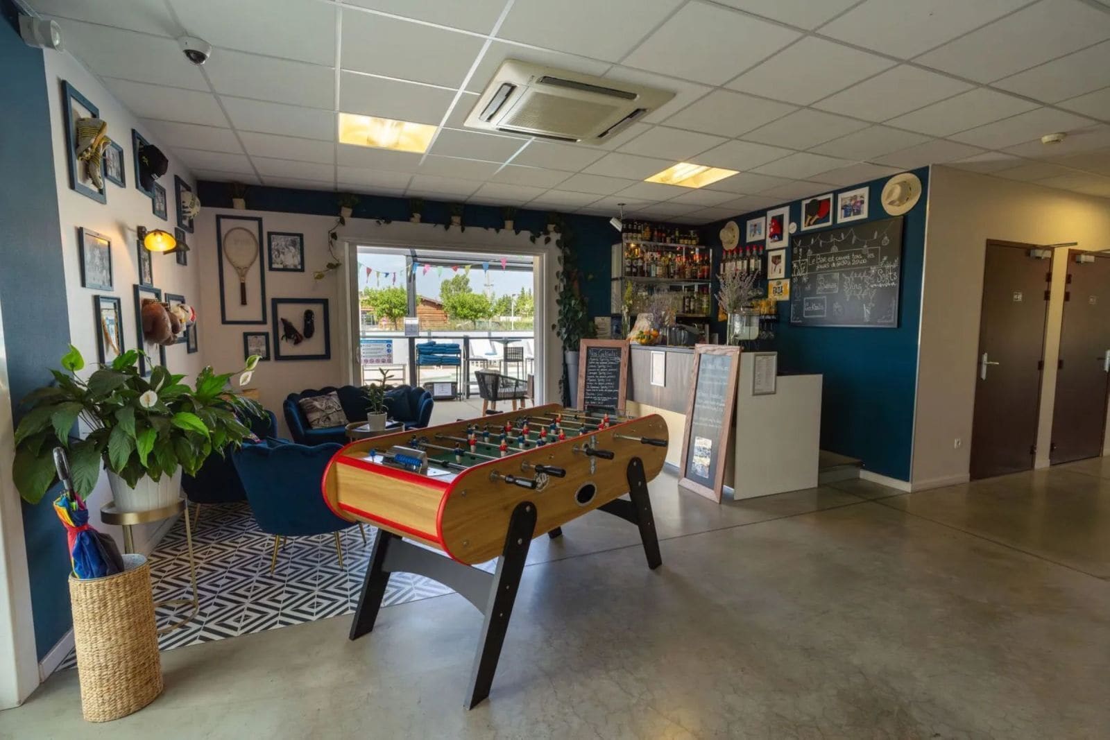 A room with a foosball table, a bar area with various bottles, and a seating area with a blue sofa and a potted plant.