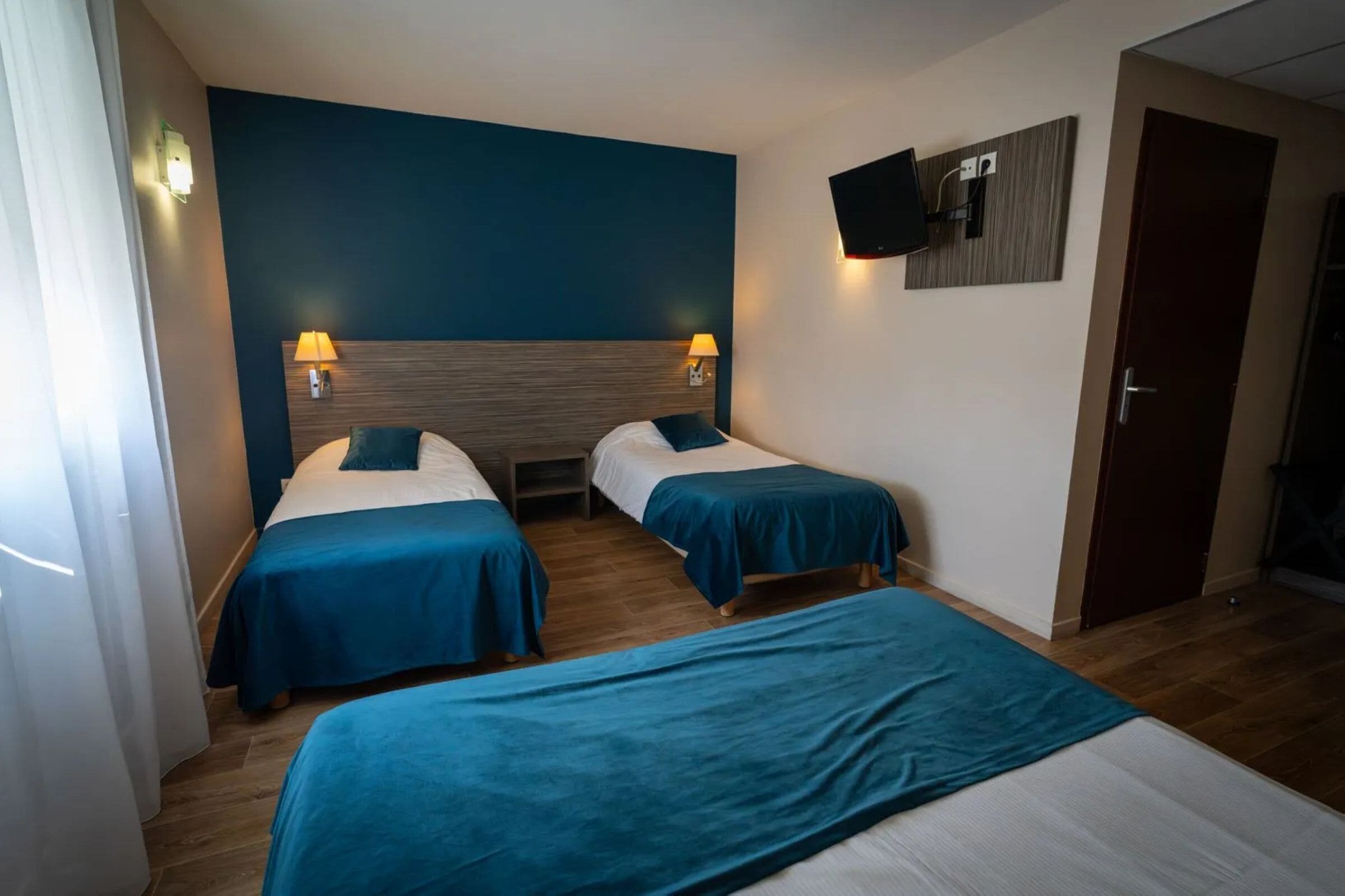 Ein Hotelzimmer mit zwei Einzelbetten, einem großen Bett, Holzböden, einem an der Wand montierten Fernseher und einer Tür.