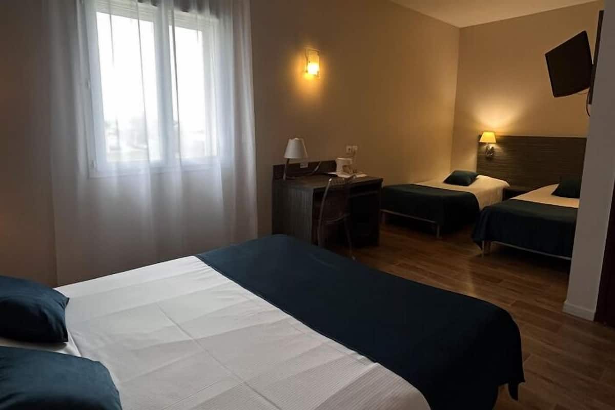 A hotel room with a large bed, two smaller beds, a desk with a chair, a TV mounted on the wall, and a window with curtains.