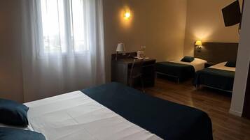Une chambre d’hôtel avec un grand lit, deux petits lits, un bureau avec une chaise, un téléviseur fixé au mur et une fenêtre avec des rideaux.