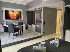 A modern hotel room with a bed, a desk, a chair, a TV, and a bathroom with a glass shower. - Luxury Room (Pescara)