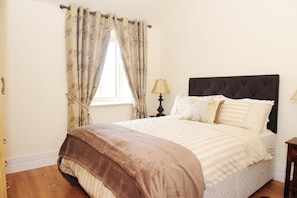 A neatly made bed with a brown headboard, a window with floral curtains, and a bedside lamp. - Sheephouse Country Courtyard (Donore)