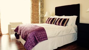 Een net opgemaakt bed met een wit en paars sprei, een accentmuur van steen en een witte stoel in een goed verlichte kamer.