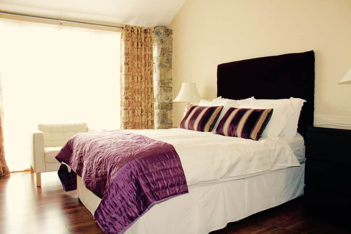 Uma cama bem arrumada com edredom branco e roxo, uma parede de pedra decorativa e uma cadeira branca em um quarto bem iluminado.