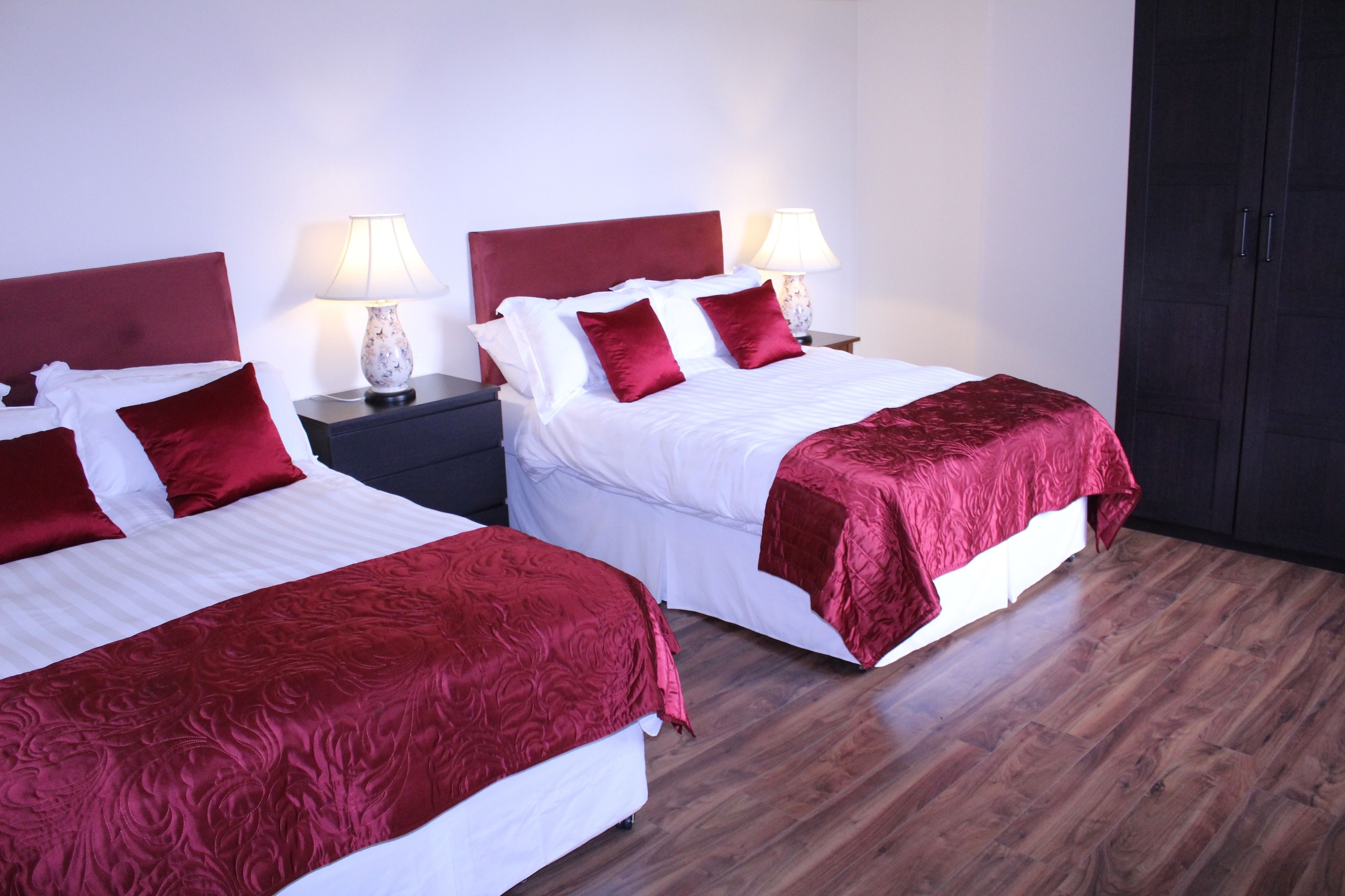 Two single beds with red headboards and white bedding, each with a bedside table and lamp. The room has wooden flooring and a dark wardrobe.