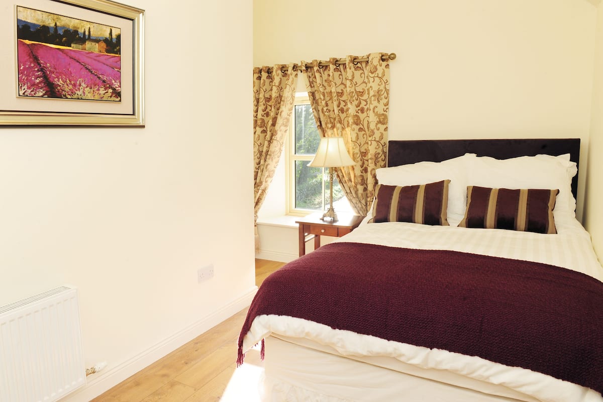 Quarto com cama, mesa de cabeceira, luminária, janela com cortinas e um quadro na parede.