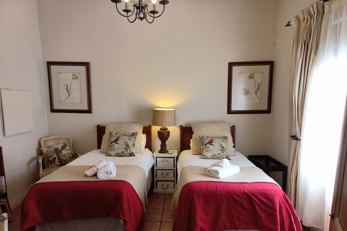 Quarto de hotel com duas camas, cada uma com colcha vermelha, mesa de cabeceira com abajur e quadros nas paredes.