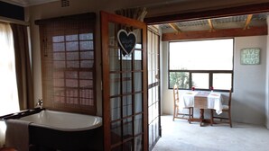 A bathroom with a bathtub, a window with blinds, and a table with chairs outside.