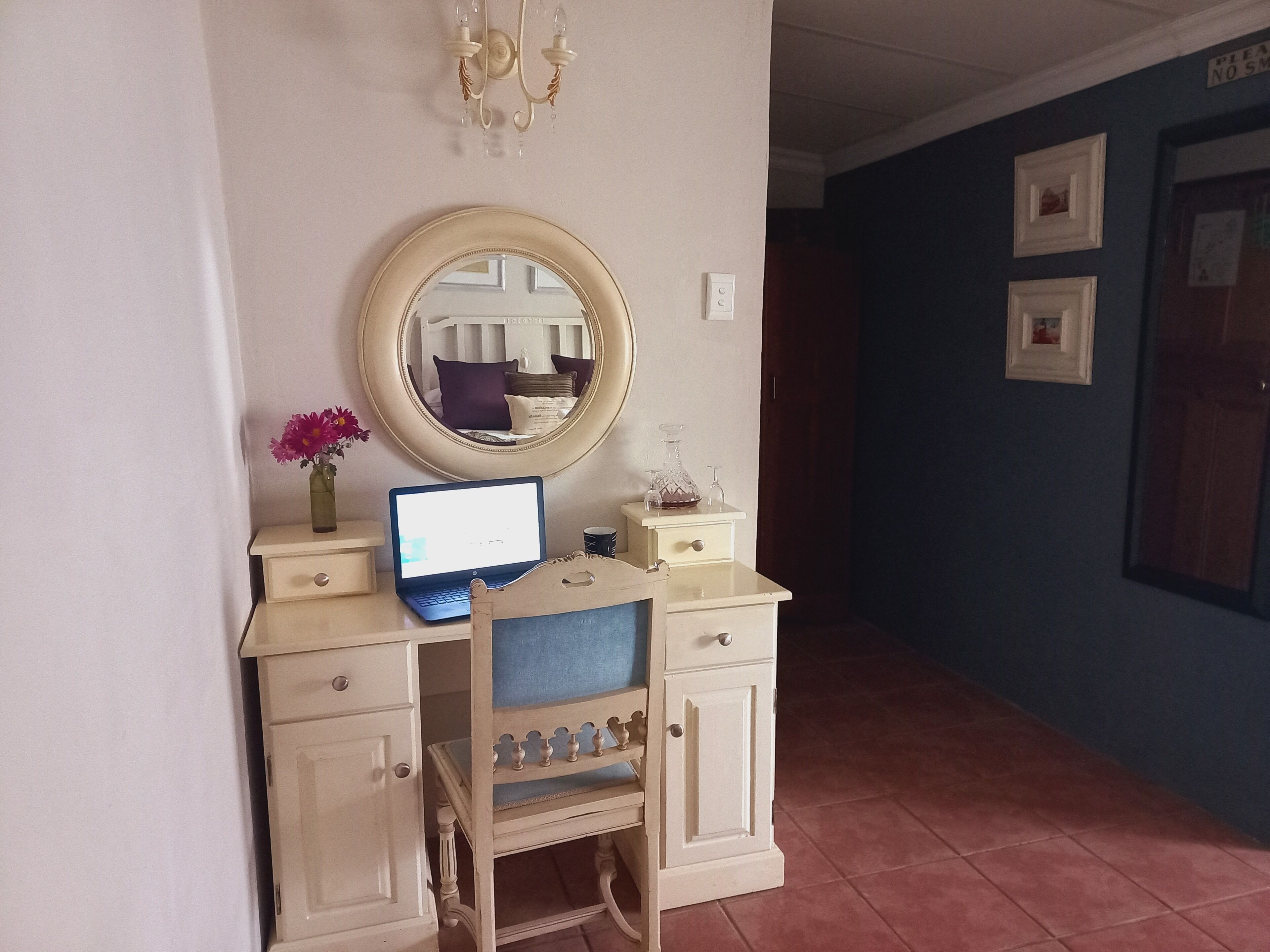 A small desk with a laptop, a mirror, and a vase with flowers.