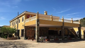 Een geel gebouw met een balkon, een bord waarop een restaurant aangegeven staat, en terrasmeubilair met stoelen en tafels.