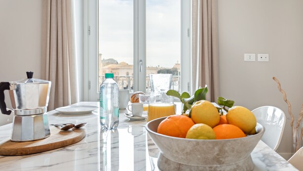Une table de petit-déjeuner avec une machine à café, une bouteille d’eau, du jus de fruits et un bol de fruits disposés sur un comptoir en marbre.