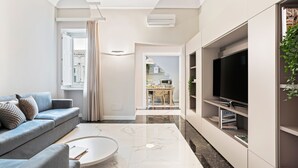 Un salon moderne avec un canapé, une table basse et un téléviseur. On peut apercevoir une cuisine à travers une porte ouverte.