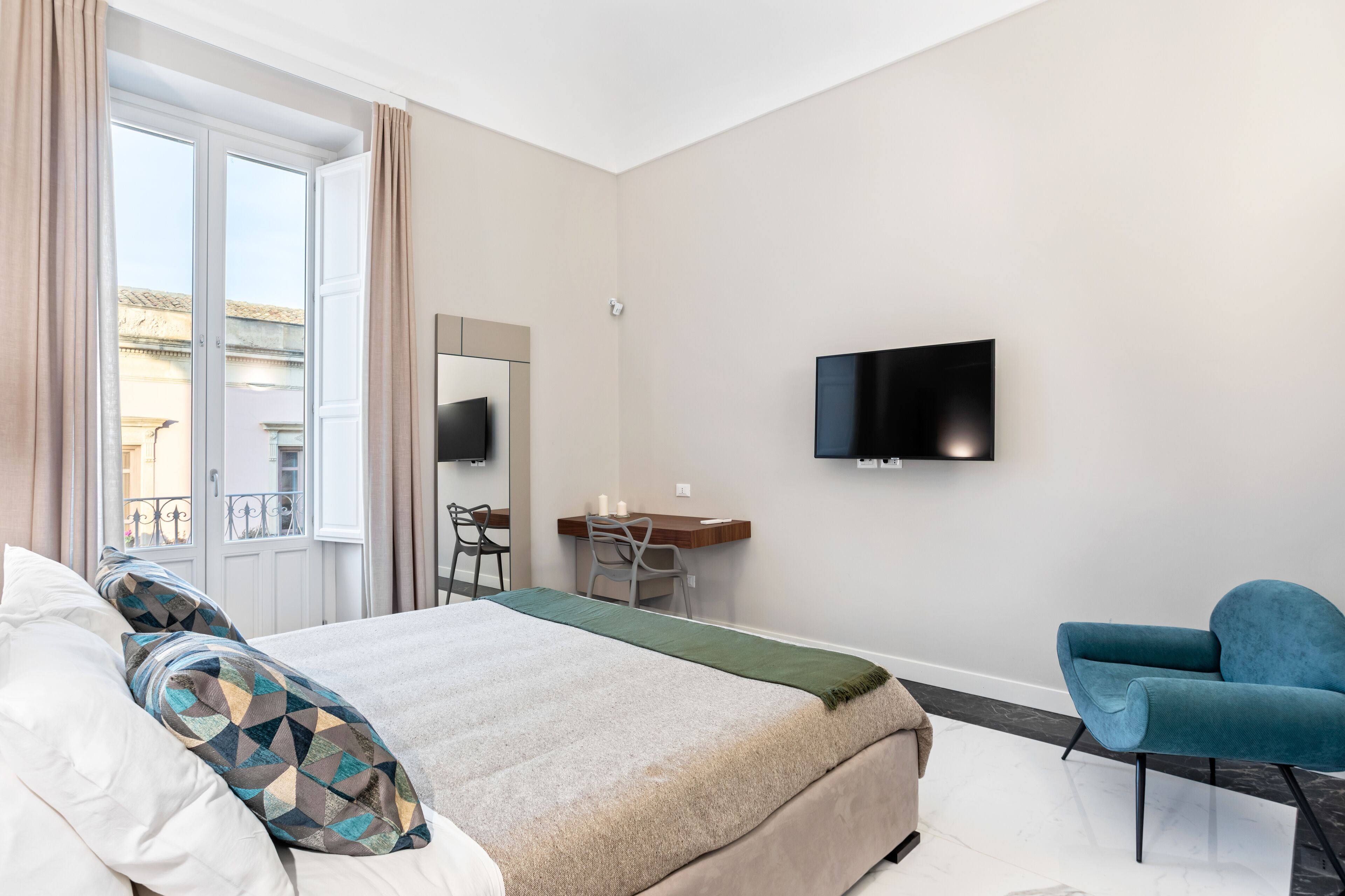 A hotel room with a bed, a desk, a chair, a TV, and a balcony with curtains.