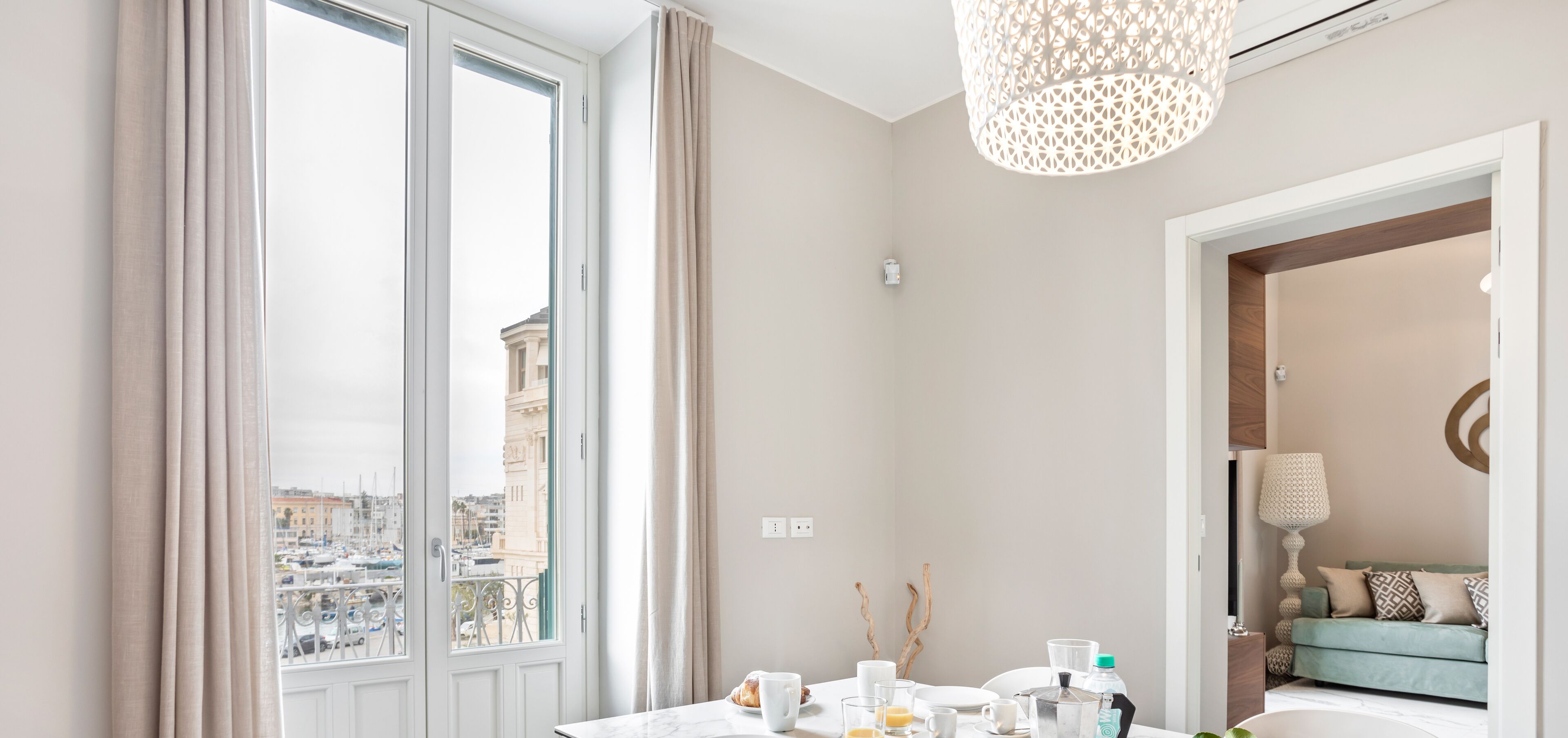 Une chambre d’hôtel moderne dotée d’une grande fenêtre, d’une table ronde avec des éléments pour le petit-déjeuner, d’une vue sur la ville et d’un coin salon confortable avec un canapé et une lampe.