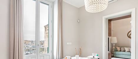 Une chambre d’hôtel moderne dotée d’une grande fenêtre, d’une table ronde avec des éléments pour le petit-déjeuner, d’une vue sur la ville et d’un coin salon confortable avec un canapé et une lampe.