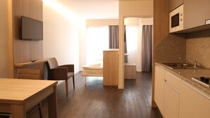 A compact hotel room with a wooden dining table, a single bed, a kitchenette with a microwave, and a television mounted on the wall. - Wego Hotel (Vuadens)