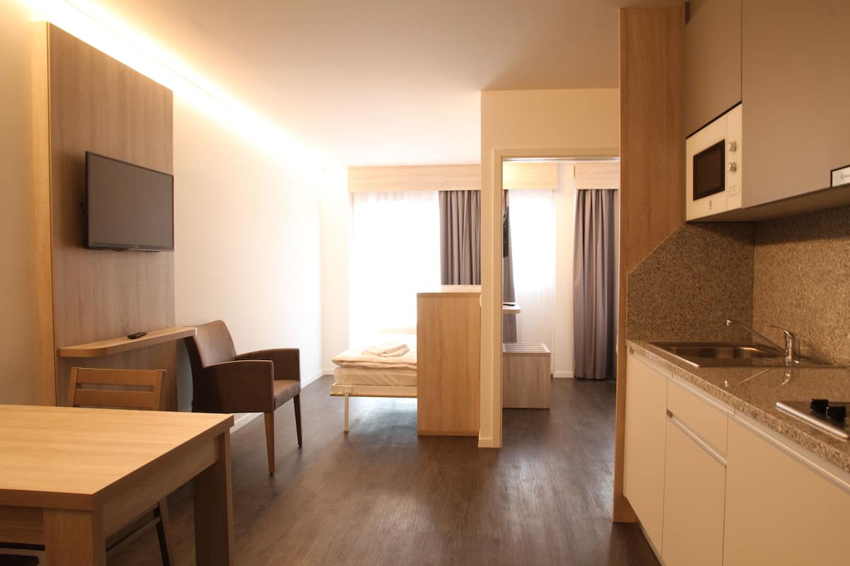 A compact hotel room with a wooden dining table, a single bed, a kitchenette with a microwave, and a television mounted on the wall.