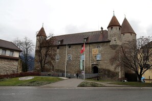A historic stone building with a central tower and two smaller towers, a flag, and a paved entrance area.