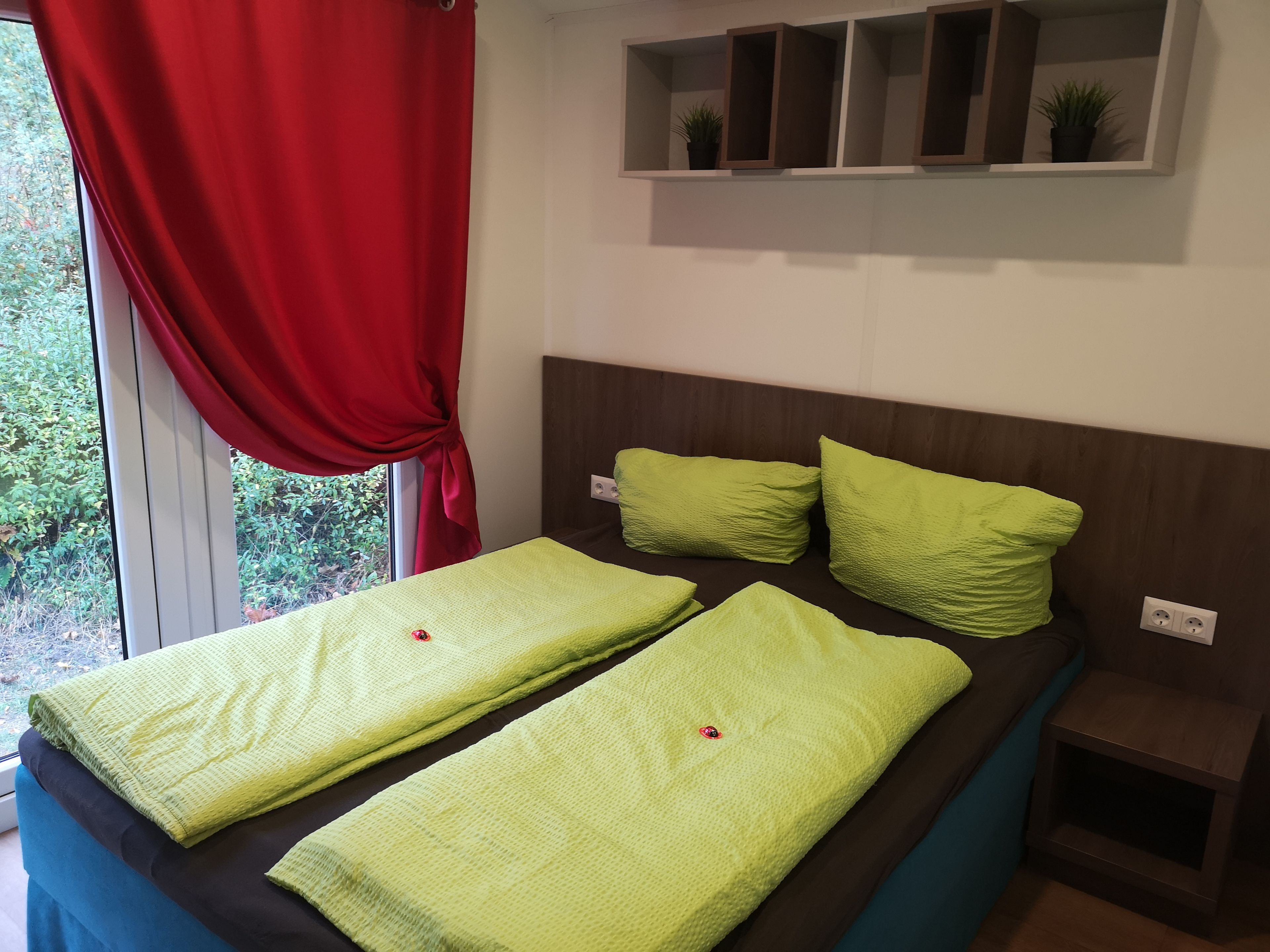 A bedroom with a bed, red curtains, a headboard, and a shelf with plants.
