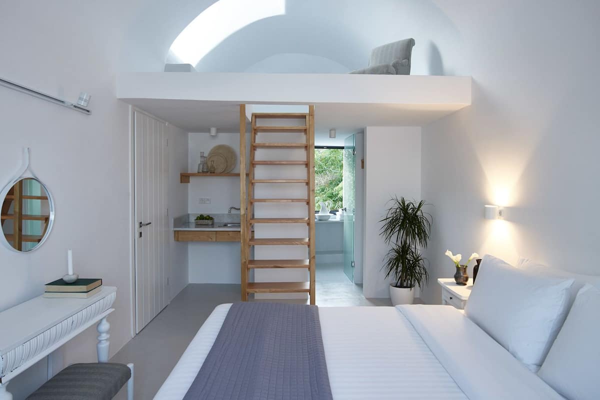A bedroom with a lofted bathroom, a wooden ladder, a bedside table, a lamp, and a potted plant.