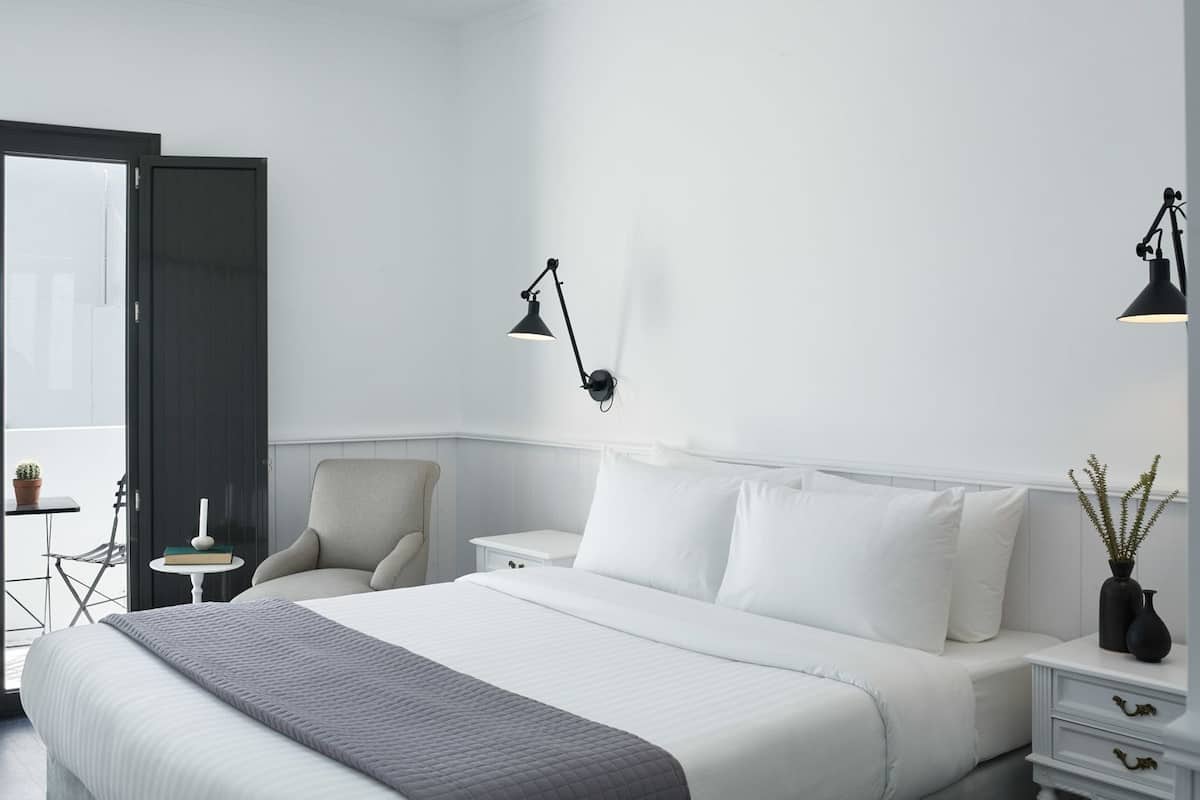 A modern hotel room with a large bed, a bedside table, a chair, and a small table with a book and a vase.