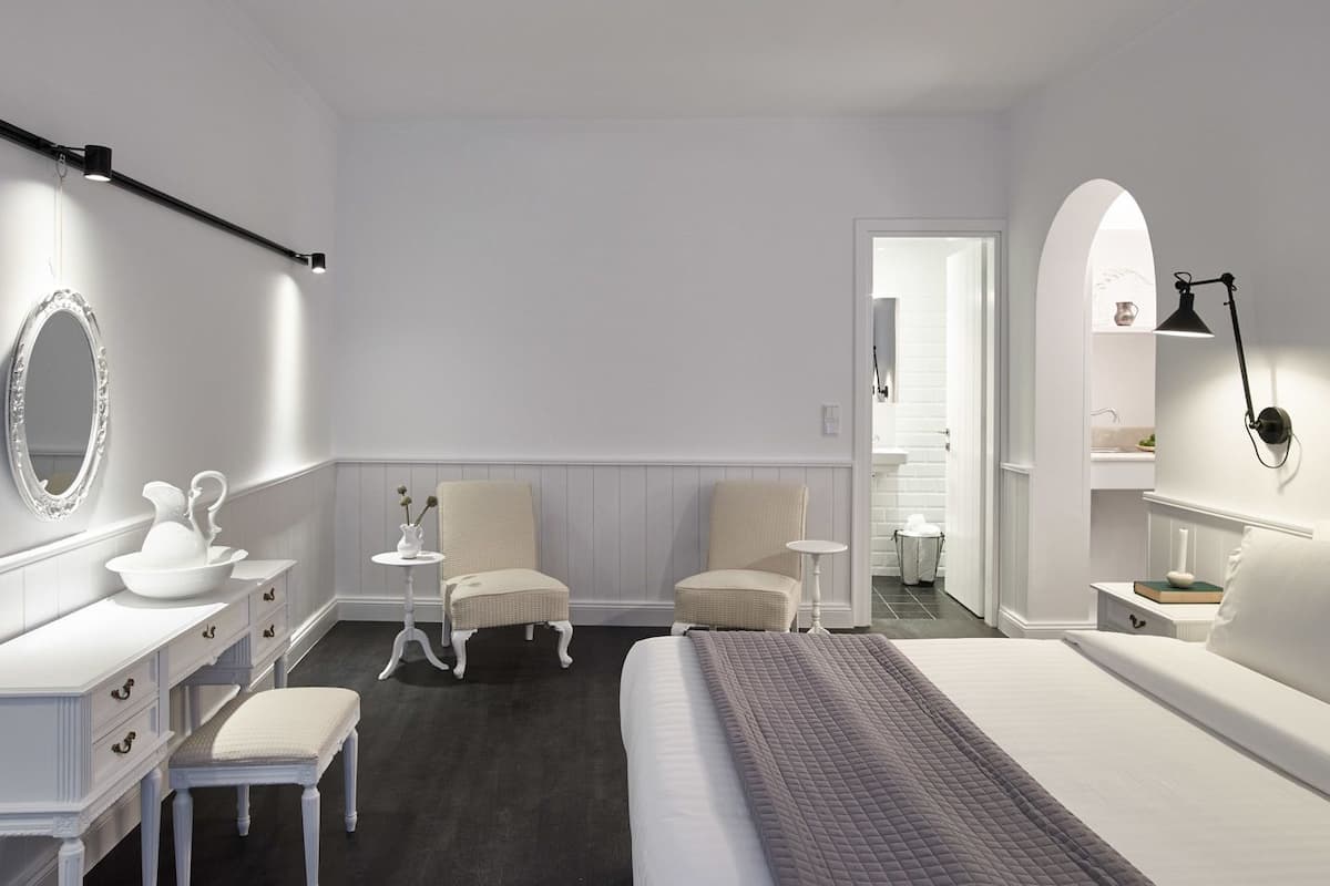 A bedroom with a bed, two chairs, a dresser, a mirror, and a nightstand with a lamp.