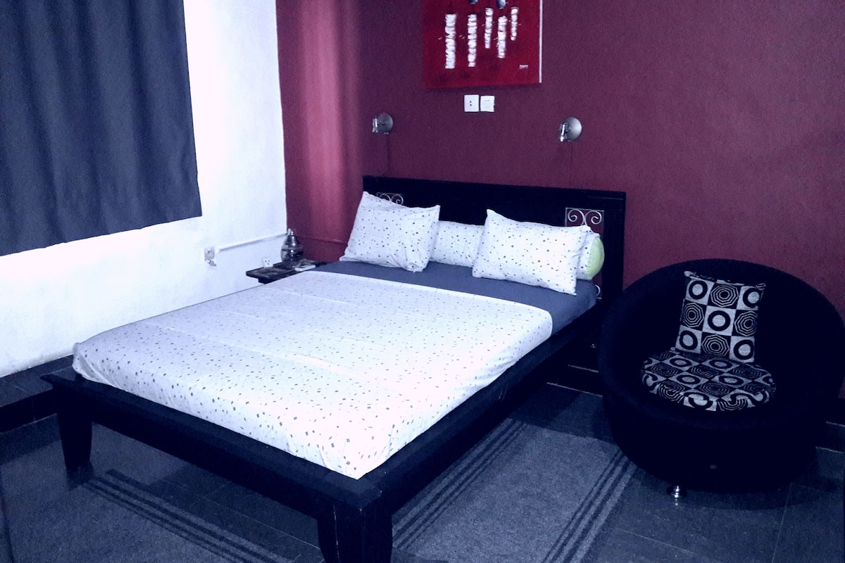 A hotel room with a bed, a chair, a red wall, and a window with curtains.