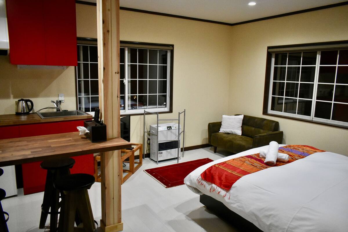 A hotel room with a bed, a desk with a chair, a kitchenette, and a window.