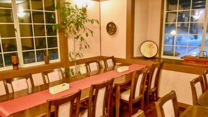 Un comedor con una mesa larga, sillas de madera, una planta en maceta y vistas al exterior a través de una ventana.