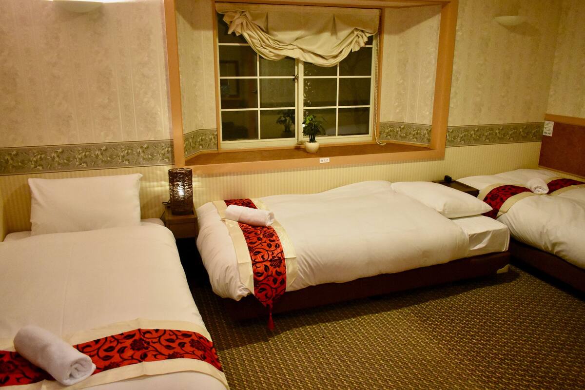 A hotel room with three single beds, each with white linens and red decorative pillows. There is a window with a curtain, a bedside table with a lamp, and a small potted plant on the windowsill.