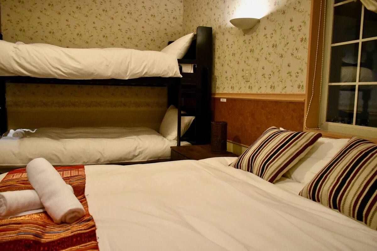 A hotel room with two beds, one with a bunk bed and the other with a standard bed. There is a wooden nightstand between the beds, a wall-mounted lamp, and a window with curtains.
