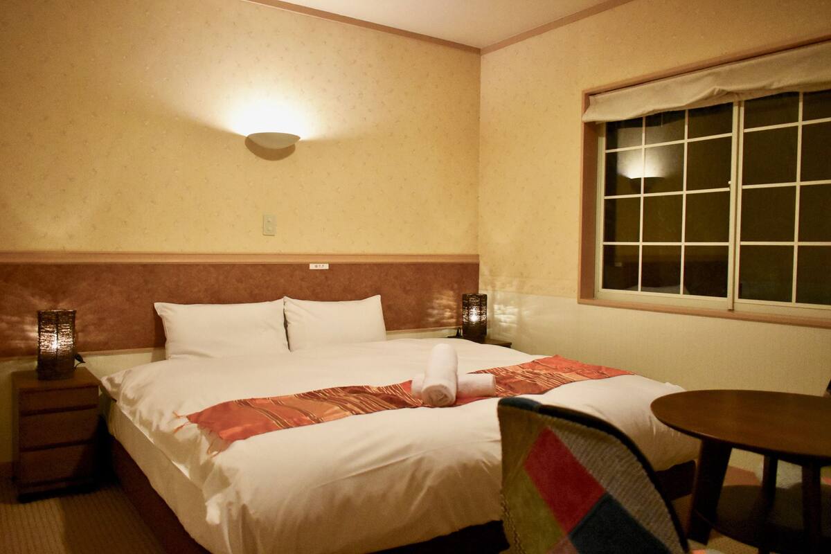 A hotel room with a neatly made bed, a bedside table with a lamp, a window with blinds, and a small round table.