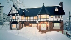 Una casa grande de dos pisos con un techo pronunciado, múltiples ventanas y una chimenea. Nieve acumulada frente a la casa.