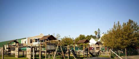 Un parque infantil con estructuras de madera, incluyendo columpios y un tobogán, rodeado de césped y árboles.