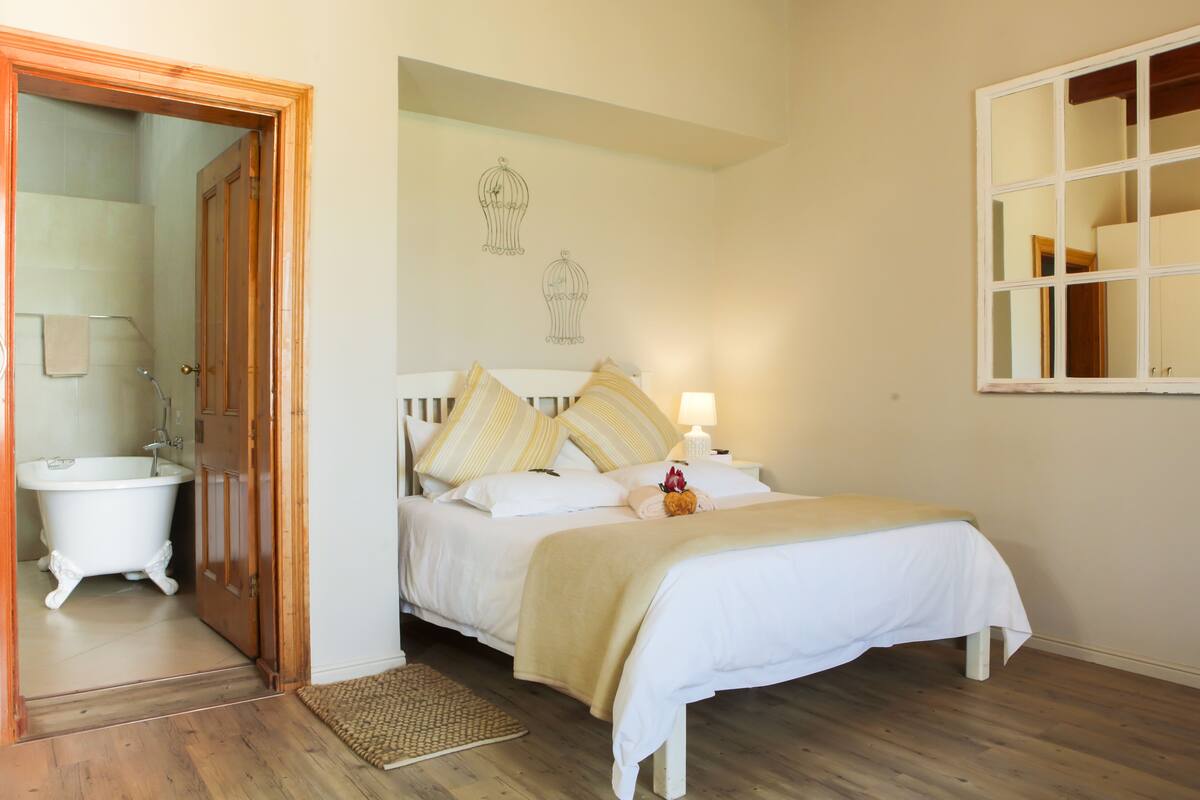 Une chambre avec un plancher en bois, un lit blanc avec des oreillers à rayures, une table de chevet avec une lampe et une porte en bois donnant sur une salle de bain avec une baignoire blanche.