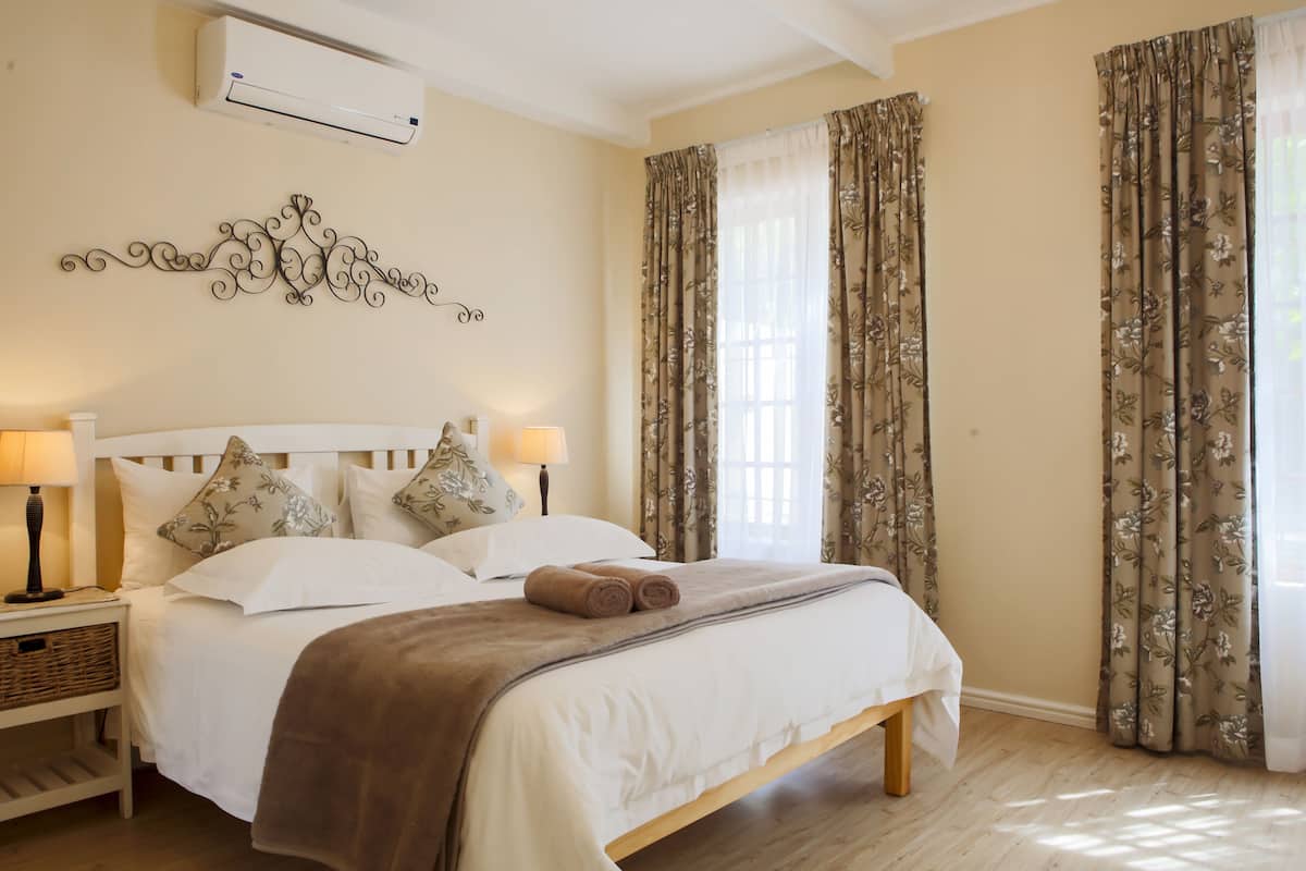 Un lit bien fait, avec une tête de lit blanche, un plaid marron et deux serviettes enroulées. On y trouve une table de chevet avec une lampe et un panier. La pièce est décorée de murs beige, d’un élément décoratif mural et de rideaux à motifs floraux.