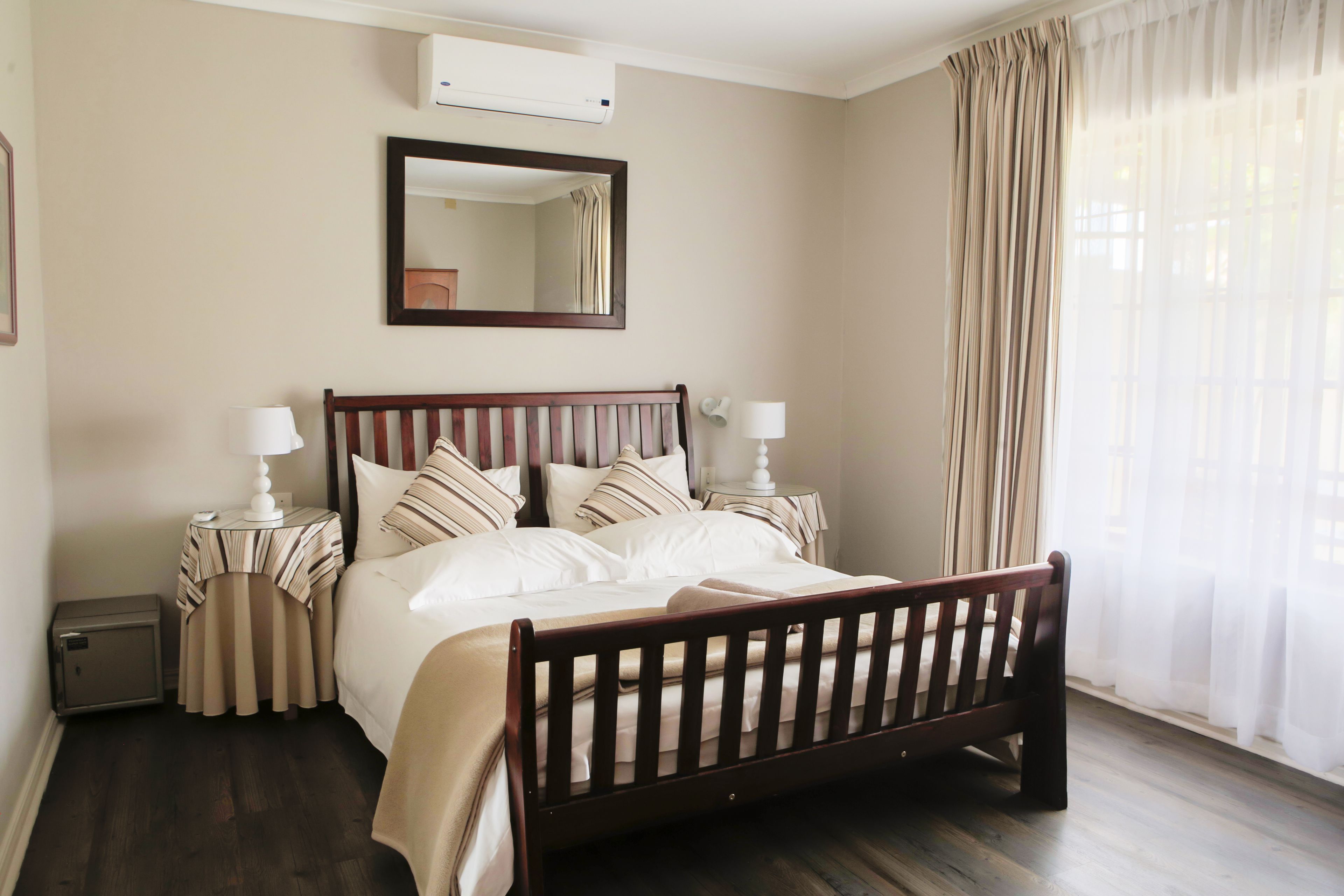 A neatly made bed with a wooden headboard, two bedside tables with lamps, a mirror above the bed, and a window with sheer curtains.