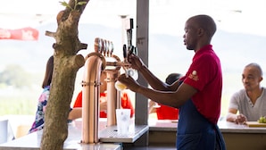 Eine Barista bereitet ein Getränk an einer Kaffeemaschine mit Messingauslauf zu, unter einer überdachten Außenbereits mit Baum und Bergblick im Hintergrund.