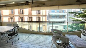 En balkon med flisegulv, fletmøbler og udsigt til en bygning med flere vinduer og balkoner.