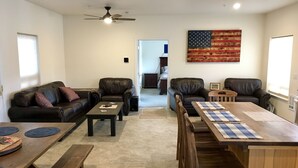 Ein Wohnzimmer mit einer Couch, zwei Sesseln, einem Couchtisch, einem Holzesstisch mit Stühlen, einem Deckenventilator und einem großen Gemälde einer amerikanischen Flagge an der Wand.