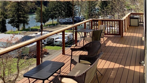 Eine Holzterrasse mit Sitzgelegenheiten im Freien, Sonnenschirmen und Blick auf einen See sowie Wohnmobile im Hintergrund.