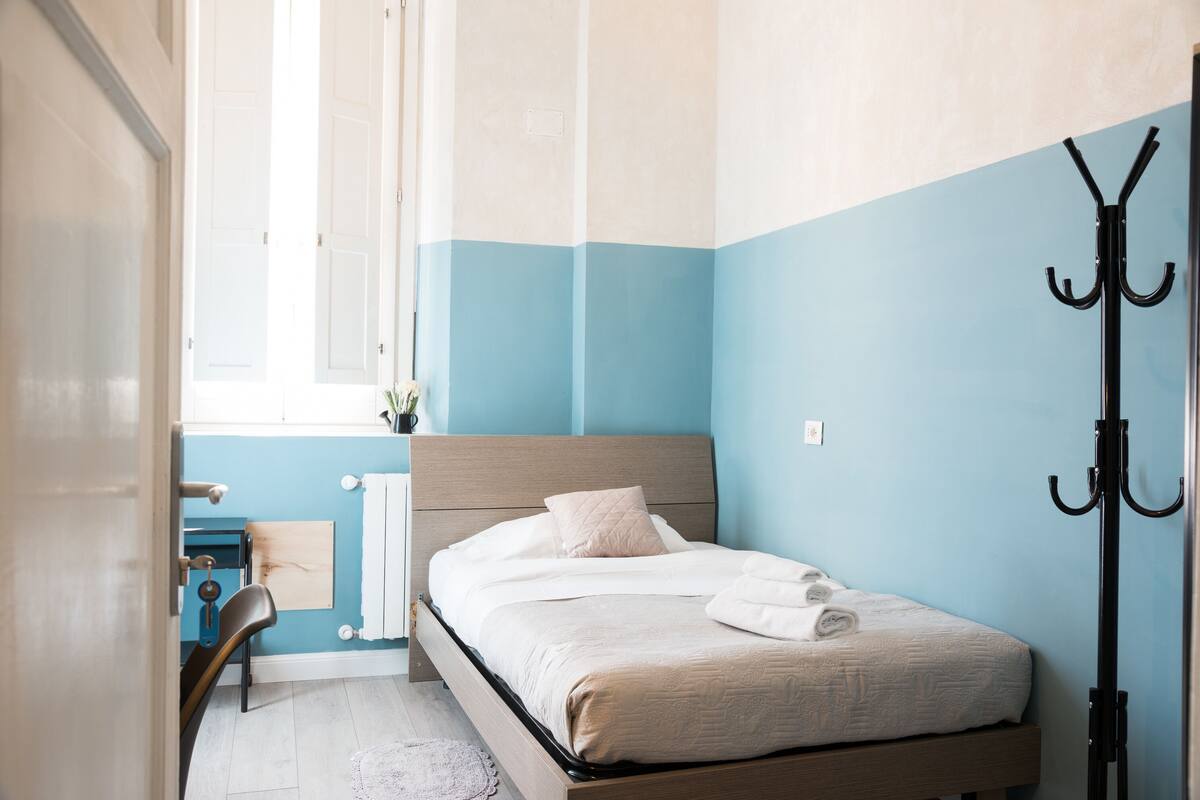 Un dormitorio con una cama, una silla, un perchero y una ventana.