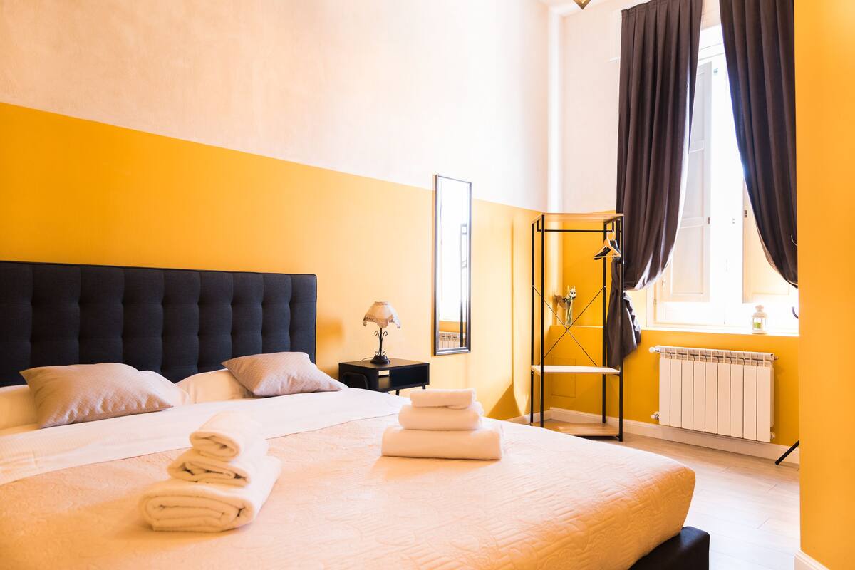 Habitación de hotel con una cama grande, una mesita de noche con lámpara, una ventana con cortinas y un radiador.