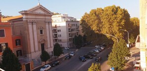 Vista de la calle con coches aparcados, un edificio de arquitectura clásica y árboles.